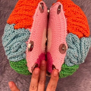 May include: A round, two-part crochet bag featuring vibrant color-blocked sections in orange, light blue, and green. The interior panels are light pink with a central white circle. Four silver-toned magnetic snap closures are visible on each side.