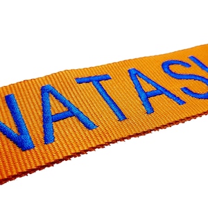Könnte beinhalten: Orangefarbener Stoffgurt mit dem Namen "NATASHA" in blau gestickt.