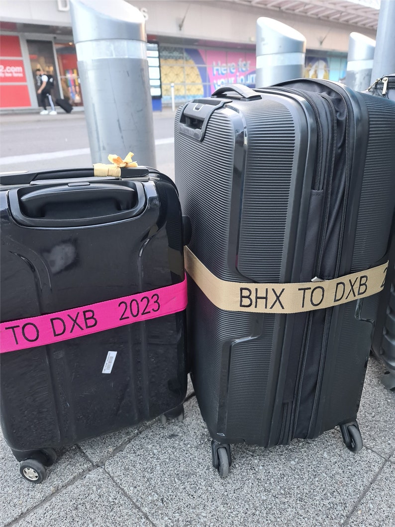 two suitcases side by side on a sidewalkat an airport. Both straps appear to be custom-made to indicate travel plans, he surroundings suggest the luggage owners are either departing from or arriving at a travel hub.