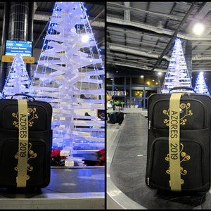 black suitcase with a personalized dijon luggage strap. The strap has text embroidered in elegant black lettering, possibly indicating a memento from a trip or event that year. The suitcase is on a baggage carousel waiting to be collected.