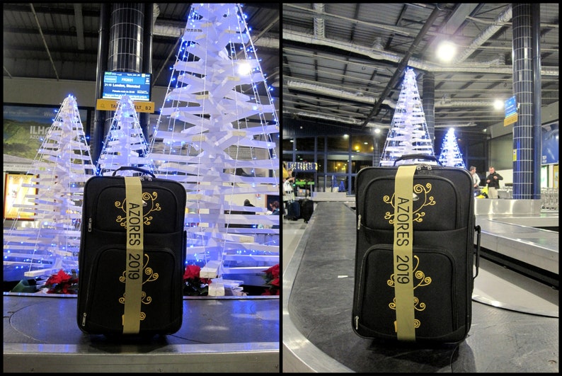 black suitcase with a personalized dijon luggage strap. The strap has text embroidered in elegant black lettering, possibly indicating a memento from a trip or event that year. The suitcase is on a baggage carousel waiting to be collected.