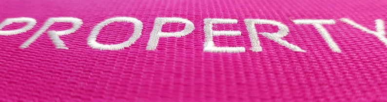 The image showcases an pink luggage strap with the name embroidered in large, white letters. The strap is bright colour enhances visibility, and the bold lettering personalizes the item, making it easier for the owner to identify their luggage.