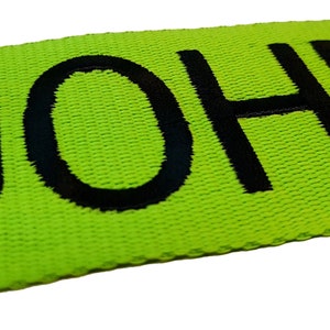 The image showcases an green luggage strap with the name embroidered in large, black letters. The strap is bright colour enhances visibility, and the bold lettering personalizes the item, making it easier for the owner to identify their luggage.