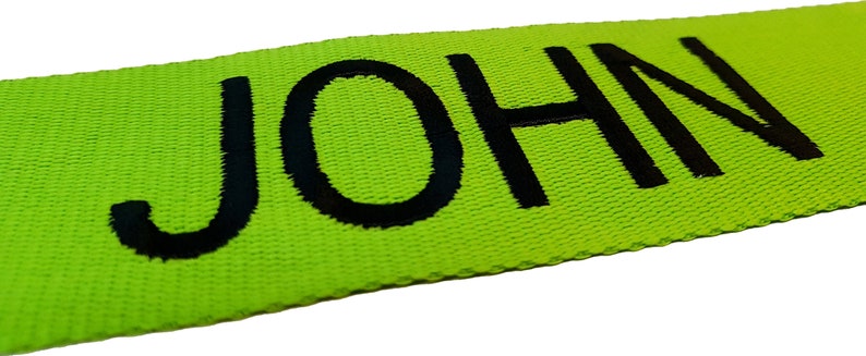 The image showcases an green luggage strap with the name embroidered in large, black letters. The strap is bright colour enhances visibility, and the bold lettering personalizes the item, making it easier for the owner to identify their luggage.