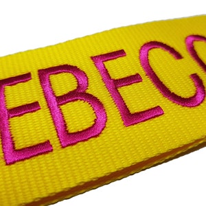 The image showcases an yellow luggage strap with the name embroidered in large, pink letters. The strap is bright colour enhances visibility, and the bold lettering personalizes the item, making it easier for the owner to identify their luggage.