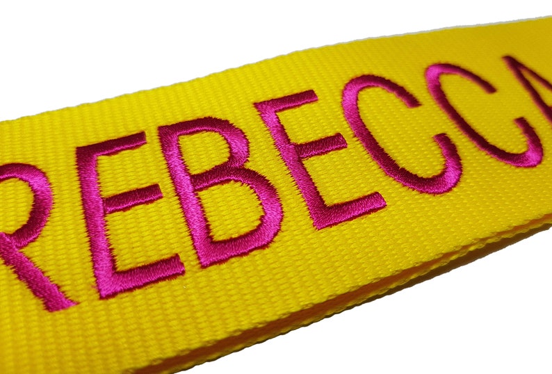 The image showcases an yellow luggage strap with the name embroidered in large, pink letters. The strap is bright colour enhances visibility, and the bold lettering personalizes the item, making it easier for the owner to identify their luggage.