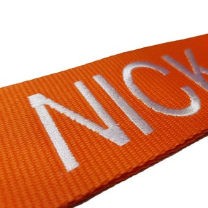 The image showcases an orange luggage strap with the name embroidered in large, white letters. The strap is bright colour enhances visibility, and the bold lettering personalizes the item, making it easier for the owner to identify their luggage.