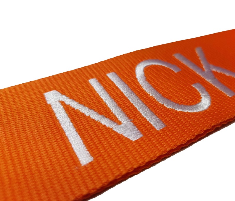 The image showcases an orange luggage strap with the name embroidered in large, white letters. The strap is bright colour enhances visibility, and the bold lettering personalizes the item, making it easier for the owner to identify their luggage.