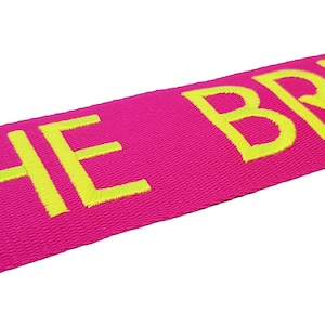The image showcases an pink luggage strap with the name embroidered in large, neon letters. The strap is bright colour enhances visibility, and the bold lettering personalizes the item, making it easier for the owner to identify their luggage.