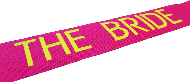 The image showcases an pink luggage strap with the name embroidered in large, neon letters. The strap is bright colour enhances visibility, and the bold lettering personalizes the item, making it easier for the owner to identify their luggage.