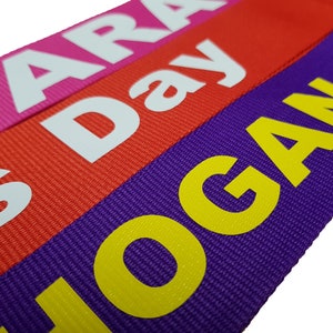 The image showcases an pink, red, purple luggage strap with the names printed. The strap is bright colour enhances visibility, and the bold lettering personalizes the item, making it easier for the owner to identify their luggage.