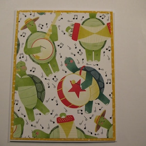 May include: Greeting card featuring a whimsical illustration of green turtles playing musical instruments. The turtles are depicted with hats and are surrounded by musical notes on a white background with a yellow border.