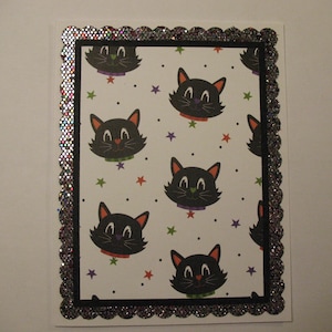 May include: A rectangular greeting card with a black cat pattern on a white background. The cats have orange ears and are wearing collars. The card is framed with a black border and a glittery, scalloped edge. Stars and dots are scattered around the cats.