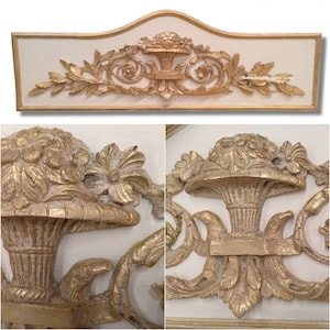 Può includere: Un pannello decorativo da parete in legno intagliato bianco e oro con un motivo floreale. Il pannello presenta un motivo centrale di un cesto di fiori con due uccelli su entrambi i lati. Il pannello è accentato con foglia d'oro e ha un bordo superiore a forma di conchiglia.