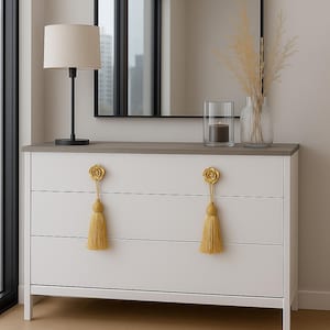 May include: White three-drawer dresser with a gray top, gold rose-shaped drawer pulls, and yellow tassels. A black-framed mirror hangs above the dresser, reflecting a window. A black lamp and decorative vase with dried plants sit on the dresser.