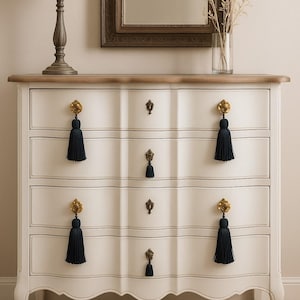 Può includere: Comò bianco con quattro cassetti, ferramenta color oro e nappe blu navy. Il comò ha un piano marrone chiaro e gambe curve. Una lampada e uno specchio sono visibili sullo sfondo, suggerendo un ambiente camera da letto.