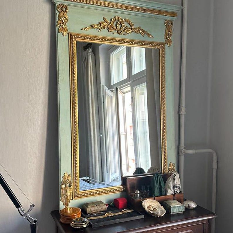 May include: A large, rectangular mirror with a mint green frame and gold accents. The mirror reflects a room with a window and curtains. The mirror is placed above a dark wooden dresser with various decorative items.