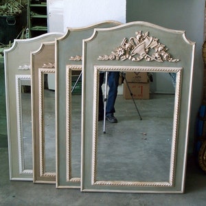 May include: Four rectangular mirrors with ornate, sage green frames and cream-coloured accents. Each mirror has a decorative crest at the top. The frames feature a rope-like trim. The mirrors are leaning against a wall.