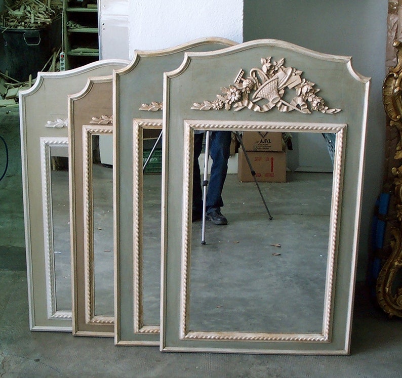 May include: Four rectangular mirrors with ornate, sage green frames and cream-coloured accents. Each mirror has a decorative crest at the top. The frames feature a rope-like trim. The mirrors are leaning against a wall.