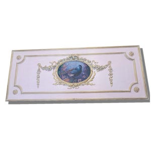 Può includere: Un pannello da parete decorativo con uno sfondo rosa chiaro e una cornice dorata. Il pannello presenta un ovale centrale con un dipinto di un uccello blu in un ambiente floreale.