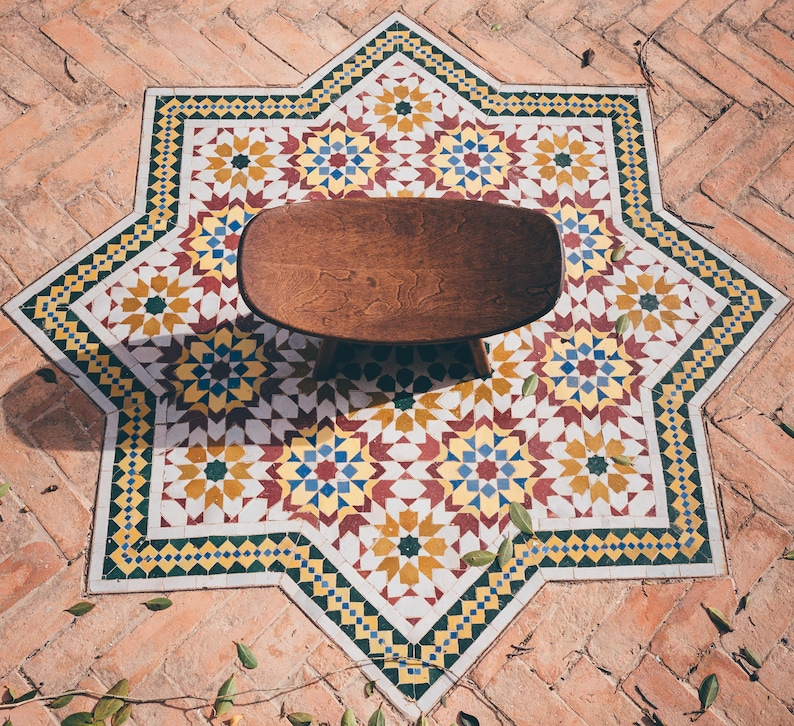 May include: A wooden stool with a rounded top sits in the center of a star-shaped tile floor. The tiles are white with yellow, blue, and red geometric patterns.