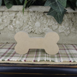 May include: A plain, unfinished wooden dog bone shape, sitting on a plaid fabric surface. The bone is light brown and has a smooth surface.