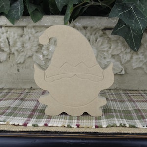 May include: Unpainted wooden cutout of a gnome with a pointy hat and a beard. The gnome is standing on a plaid surface.