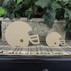 May include: Two wooden football helmet shapes, one larger than the other, are on a plaid surface. The helmets are unpainted and ready for crafting.