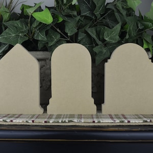 May include: Four unfinished wooden grave-shaped signs with rounded tops. The signs are beige and have a smooth surface. They are arranged in a row on a brown surface.