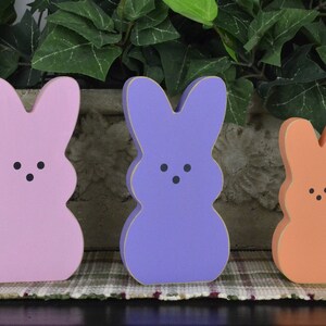 May include: Three wooden bunny figurines painted in pastel pink, purple, and orange. The bunnies have black eyes and a simple, rounded shape.