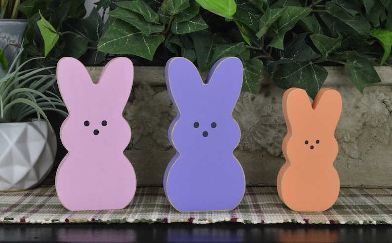 May include: Three wooden bunny figurines painted in pastel pink, purple, and orange. The bunnies have black eyes and a simple, rounded shape.