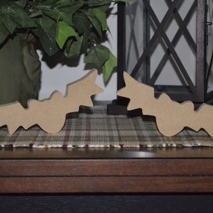May include: Two wooden bat cutouts, painted brown, are displayed on a plaid surface. The bats are facing each other, with their wings spread out.