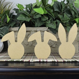 May include: Three unfinished wooden bunny shapes, standing in a row on a plaid surface. The bunnies are all the same size and shape, but have different ear styles.