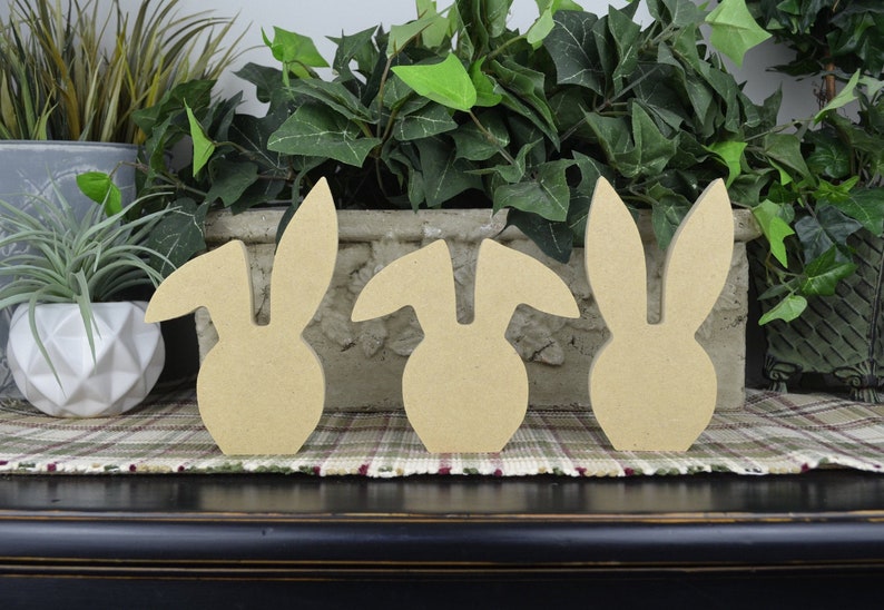 May include: Three unfinished wooden bunny shapes, standing in a row on a plaid surface. The bunnies are all the same size and shape, but have different ear styles.