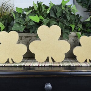 May include: Three wooden shamrock shapes in different sizes, painted in a light brown color. The shamrocks are arranged in a row on a plaid surface.
