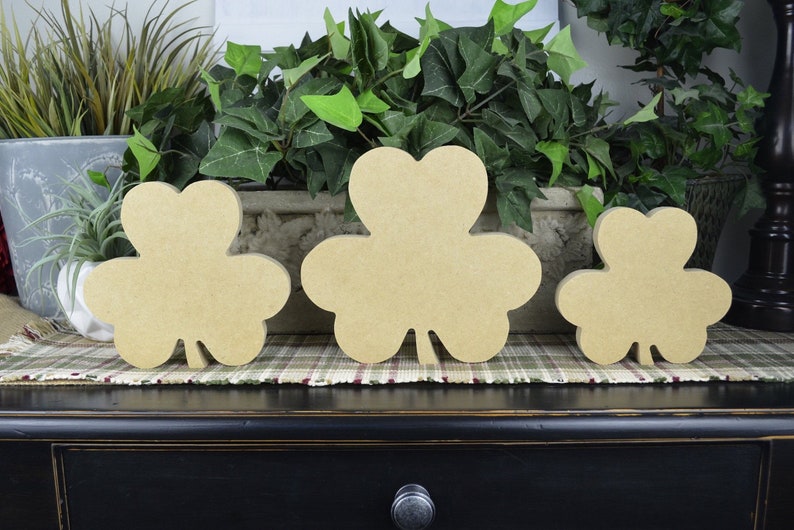May include: Three wooden shamrock shapes in different sizes, painted in a light brown color. The shamrocks are arranged in a row on a plaid surface.