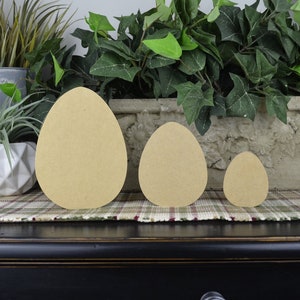 May include: Three wooden eggs in different sizes, painted in a light brown color. The eggs are arranged in a row from largest to smallest on a plaid surface.