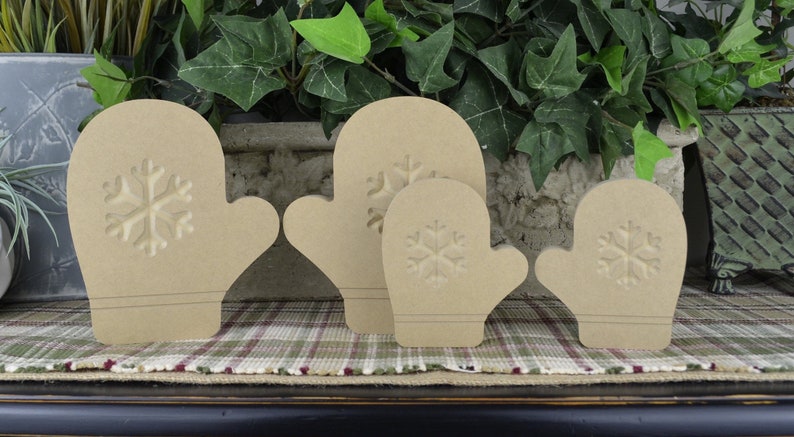 K&ouml;nnte beinhalten: Vier unfertige Holz-Handschuh-Ornamente mit Schneeflocken-Design. Die Handschuhe sind in verschiedenen Gr&ouml;&szlig;en und in einer Reihe auf einer karierten Oberfl&auml;che angeordnet.