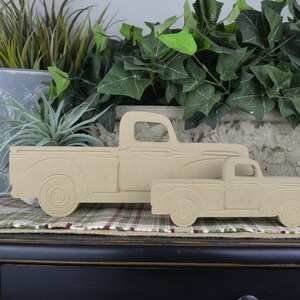 May include: Two wooden cutout trucks, one larger than the other, are displayed on a plaid surface. The trucks are a light brown color and have a simple design.