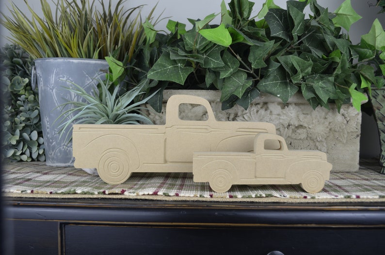 May include: Two wooden cutout trucks, one larger than the other, are displayed on a plaid surface. The trucks are a light brown color and have a simple design.