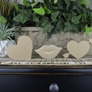 Op de afbeelding: Drie uitgesneden houten vormen: een hart, een hart met een pijl en een paar lippen. De vormen zijn allemaal ongeverfd en gemaakt van natuurlijk hout.