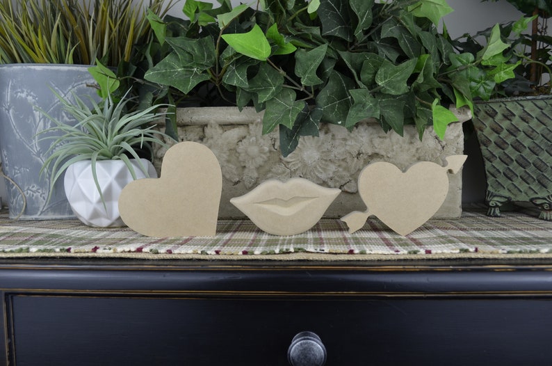 Op de afbeelding: Drie uitgesneden houten vormen: een hart, een hart met een pijl en een paar lippen. De vormen zijn allemaal ongeverfd en gemaakt van natuurlijk hout.