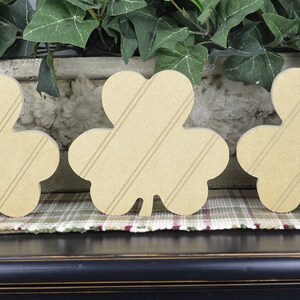 May include: Three wooden shamrock shapes with a striped pattern. The shamrocks are light brown and are arranged in a row on a plaid surface.