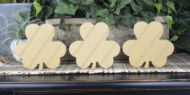 May include: Three wooden shamrock shapes with a striped pattern. The shamrocks are light brown and are arranged in a row on a plaid surface.