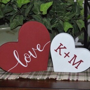 Puede incluir: Dos letreros de madera en forma de coraz&oacute;n. Uno es rojo con la palabra "love" escrita en blanco. El otro es blanco con las iniciales "K+M" escritas en rojo.