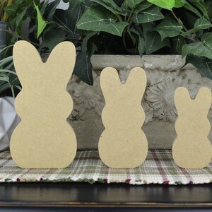 May include: Three unfinished wooden bunny shapes in different sizes. The bunnies are standing on a plaid surface.