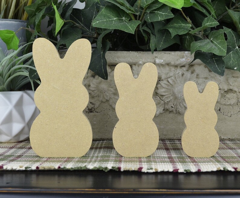 May include: Three unfinished wooden bunny shapes in different sizes. The bunnies are standing on a plaid surface.