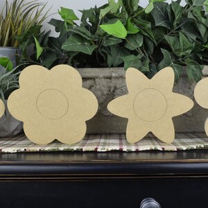 May include: Four unfinished wooden flower shapes, each with a circular cutout in the center. The flowers are arranged in a row on a dark wooden surface.