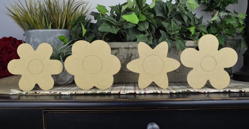 May include: Four unfinished wooden flower shapes, each with a circular cutout in the center. The flowers are arranged in a row on a dark wooden surface.