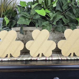 May include: Three wooden shamrock shapes with a wood grain pattern. The shamrocks are light brown and are arranged in a row on a table.